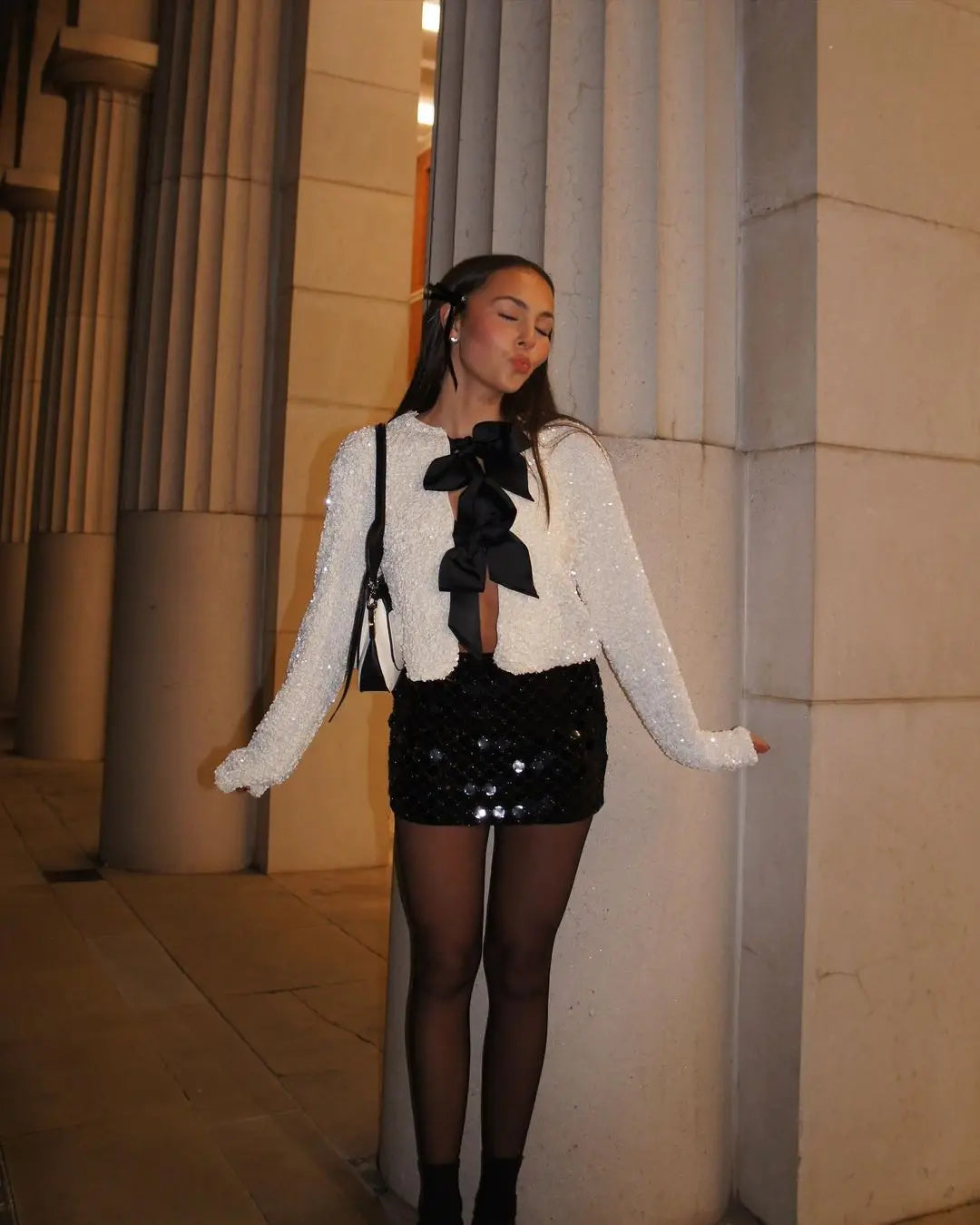 Woman in a stylish outfit with a white jacket and black sequin skirt standing against a stone wall.