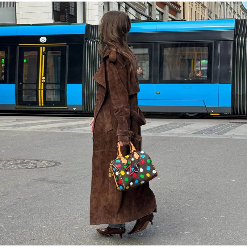 Person in a long brown coat holding a colorful polka dot bag on a city street with a blue bus in the background.