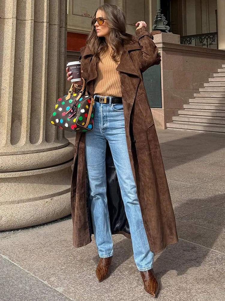 Woman in a brown coat and jeans holding a colorful polka dot bag and coffee cup, standing outdoors.