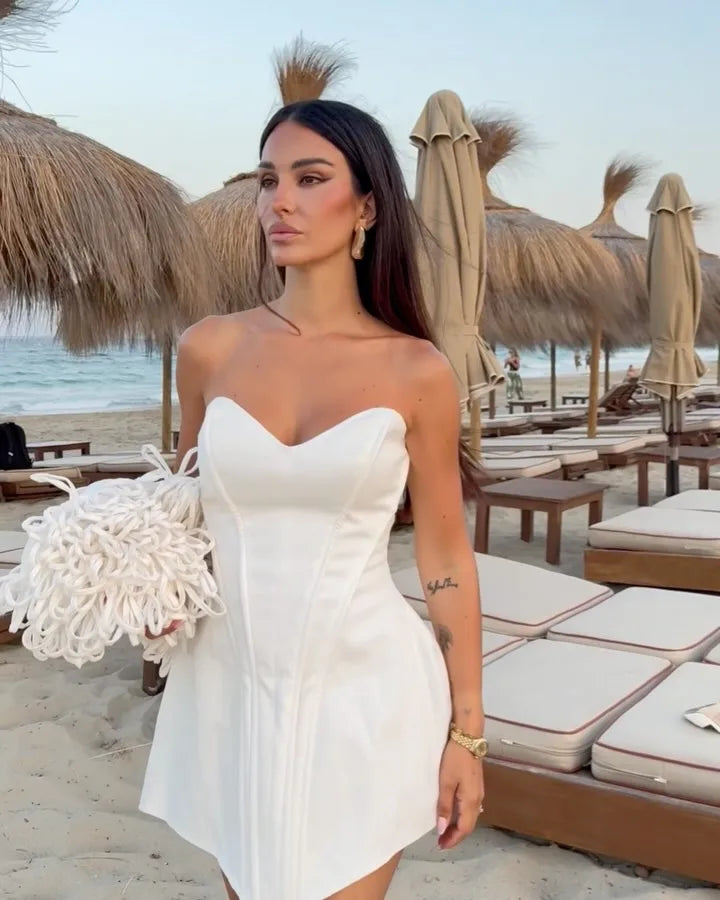 Woman in a white dress holding a bouquet on a beach with umbrellas and lounge chairs.