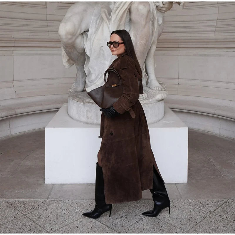 Woman in a brown coat and black boots standing in front of classical statues.