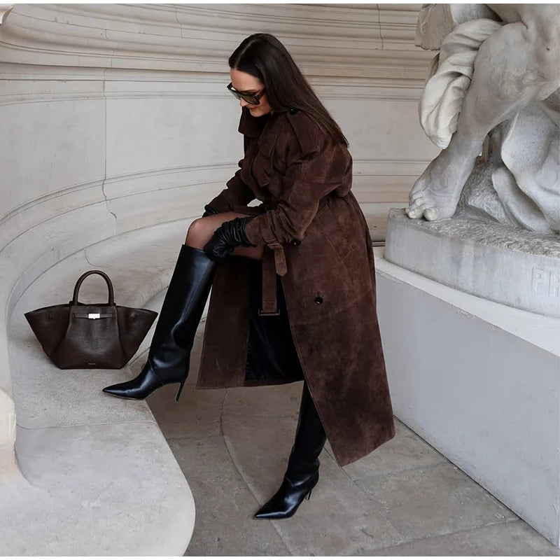 Woman in a brown coat and black boots standing next to a statue with a handbag on a stone bench.
