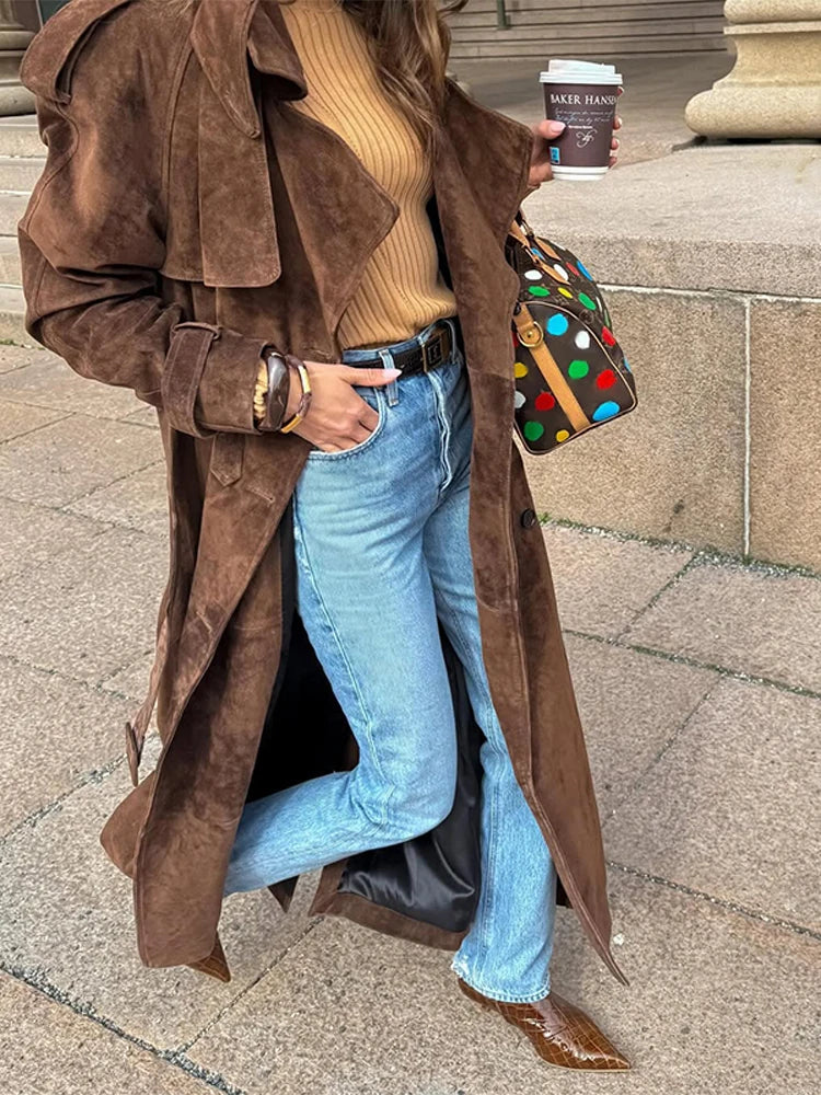 Person wearing a brown suede coat, beige sweater, blue jeans, and holding a colorful polka dot handbag.